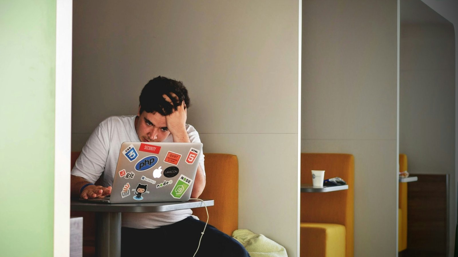 A man in a white shirt sitting at a coffee shop table, holding his head in frustration while looking at a MacBook
