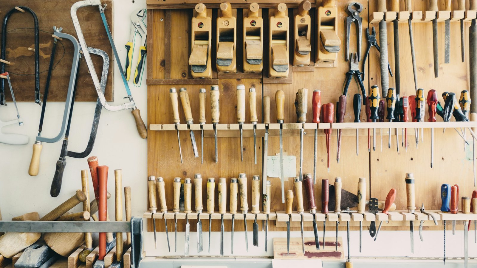 Assorted well-worn hand tools hanging in a wooden tool rack on a workshop wall