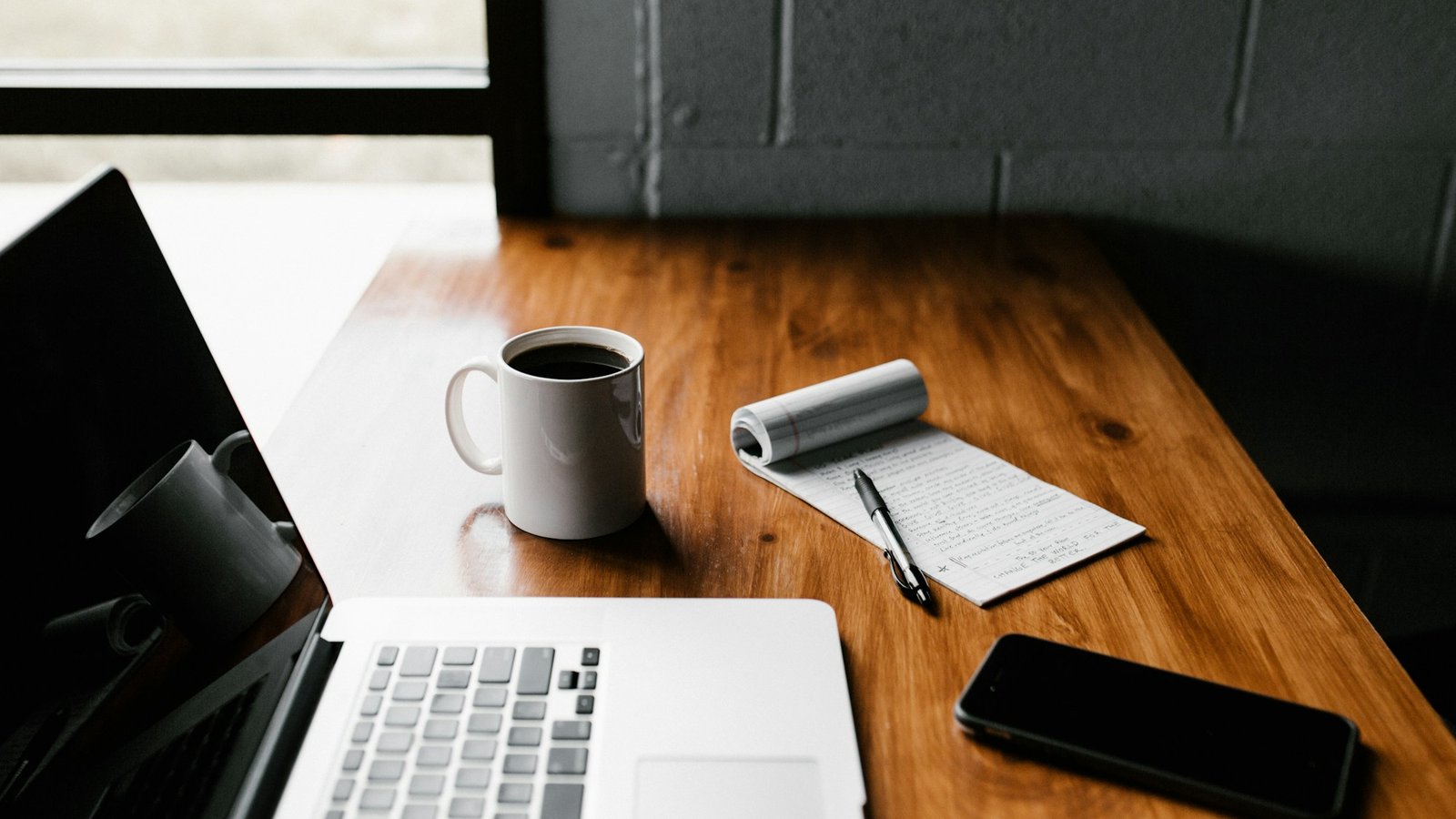 A MacBook, white ceramic mug, and smartphone arranged on a wooden desk