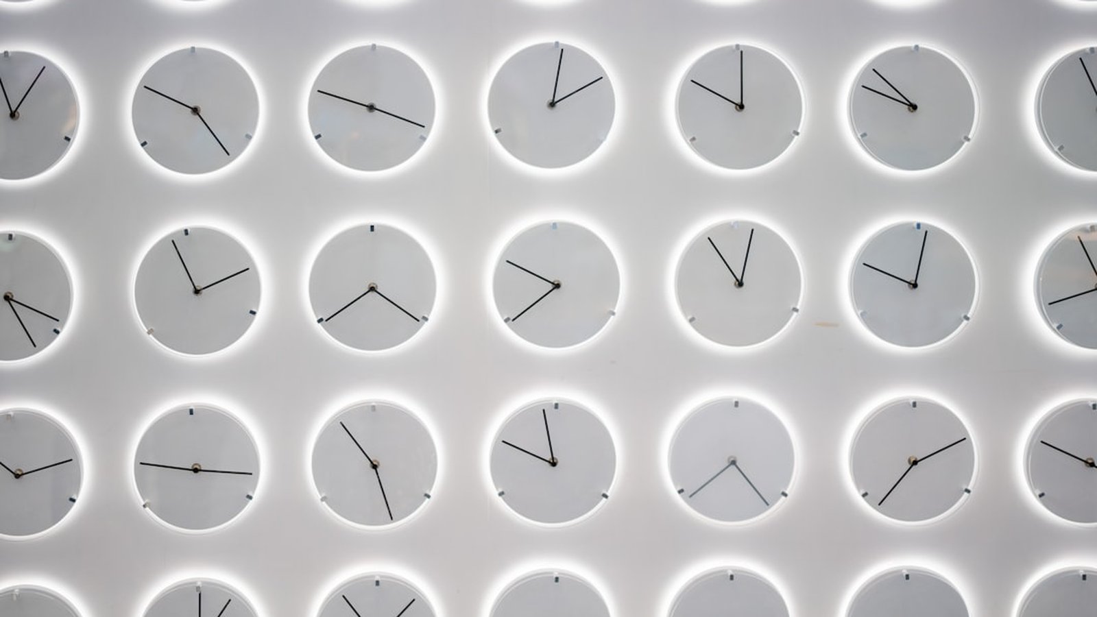 A white wall covered with many analog clocks showing different times
