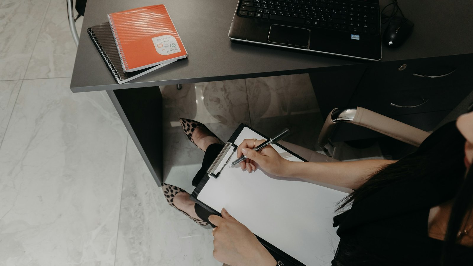 A person writing on a clipboard at a desk next to an open laptop